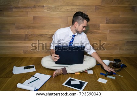 A barefoot businessman sitting on the floor against a wall and using laptop and other gadgets. Young man works at home in funny clothes and start up project, remote working.
