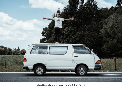 Barefoot Caucasian Young Man Dressed In A T-shirt And Black Trousers Standing With His Arms Crossed And Feet Together On The Roof Of An Old White Van, Rotorua, New Zealand