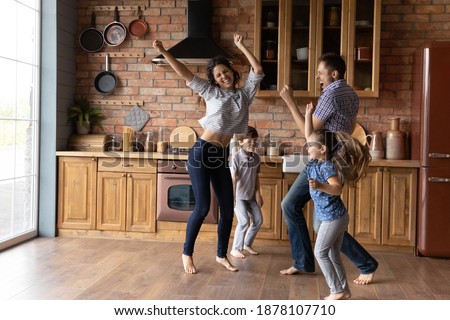Barefoot dances. Overjoyed millennial family with little daughter son having fun listening to music dancing. Active mom dad and two junior school age kids jump at kitchen celebrate moving to new home