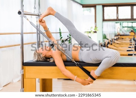 Barefoot female raising and bending legs while lying on pilates cadillac and stretching quadriceps during training in light studio