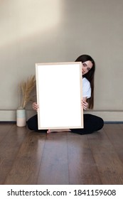 Barefoot girl with black hair in white t-shirt sitting on brown wooden floor and holding light wooden mockup frame, vase with beige pampas grass