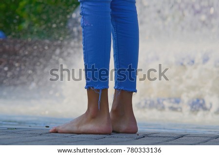 Barefoot girl standing at a swimming pool. Close up legs and feets of young woman in blue jeans standing with splashing water on background.