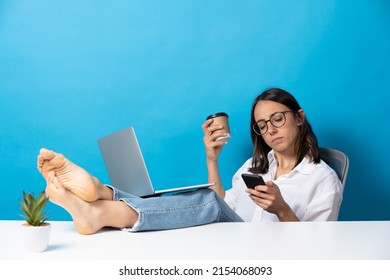 Barefoot Hispanic Woman Relaxing And Working While Sitting Feet On Desk Isolated On Blue Background. Office Worker Chatting On Phone.