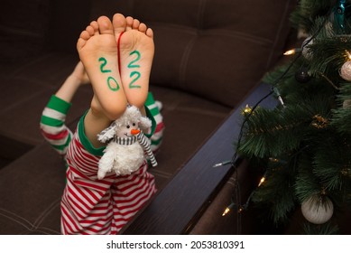 barefoot kid in striped pants lies upside down on the sofa near the Christmas tree. The numbers 2022 are painted on the feet. Cozy, cheerful, festive atmosphere. Santa's helper. Fun childhood