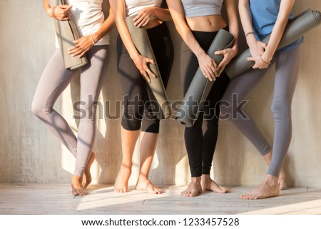 Barefoot legs feets of sportive young caucasian women in sportswear. Girls standing in a row near wall holding yoga mats rubber carpets, four females before or after sports workout at fitness studio