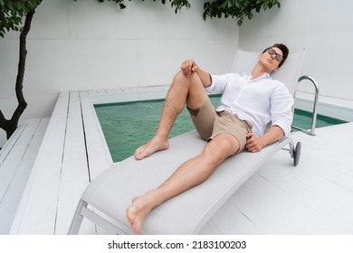 Barefoot Man In Shorts And Eyeglasses Lying On Deck Chair Near Pool