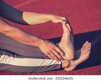 Barefoot mature woman stretching tendons and ankles on red running track in evening sun. Middle aged body in long leggings - Shutterstock ID 2101246255
