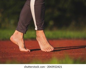 Barefoot mature woman walks on tiptoe,   stretching tendons and ankles on red running track in evening sun. Middle aged body in long leggings
