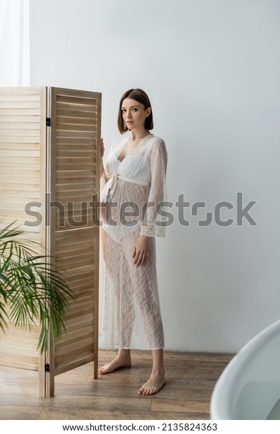 Barefoot pregnant woman in robe standing near\
folding screen in\
bathroom