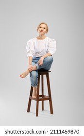 Barefoot Senior Woman In Jeans And White Blouse Sitting On Tall Chair In Studio Throws One Leg