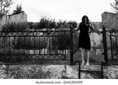 A barefoot woman in a black dress stands on a metal fence near an abandoned concrete structure