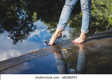 Barefoot Woman In Blue Jeans Standing On A Pavement By The Water, Dipping Toe In Water