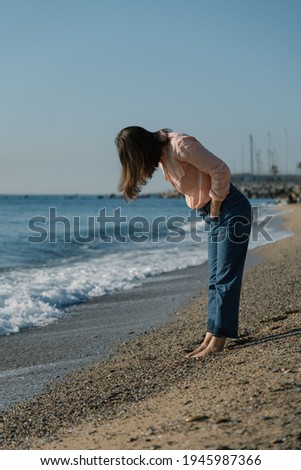 Barefoot woman in pink shirt and blue jeans looks down at her feet on beach.