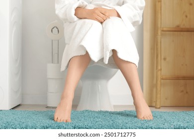 Barefoot woman sitting on toilet bowl in restroom, closeup