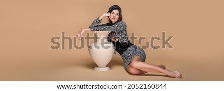 barefoot woman in zebra print outfit and black beret posing near clay vase on beige, banner