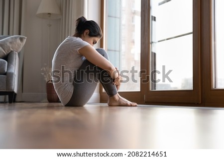 Barefoot young indian woman sitting on warm floor embracing knees, stretching back muscles after workout. Stressed unhappy lady feeling depressed, thinking of personal problems at home.