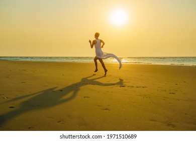 Barefoot Young Woman In White Clothes Run Away On The Sand On A Deserted Beach At Sunset Over The Sea. Long Skirt Flutters In The Wind. Freedom And Travel. Anti Lockdown Covid