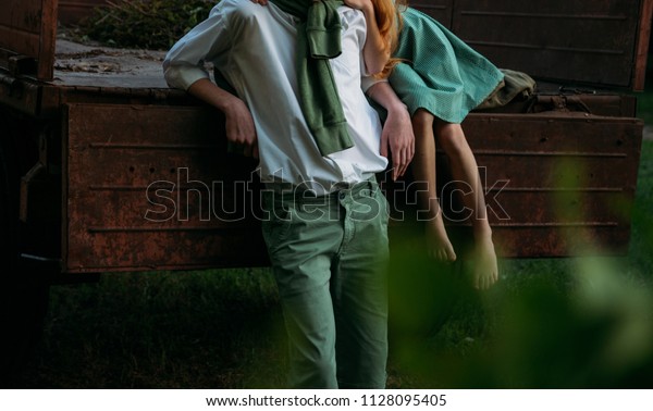 a barefooted girl in a green, vintage dress, the\
dress sits on the back of a red, old car and hugs the barefoot\
neck, next to a standing guy who is dressed in a white shirt, green\
shorts with a green
