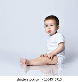 Barefooted Infant Baby Boy Toddler In White Bodysuit Is Sitting Sideways On The Floor Holding Hands Knees Looking Forward On A Gray Background