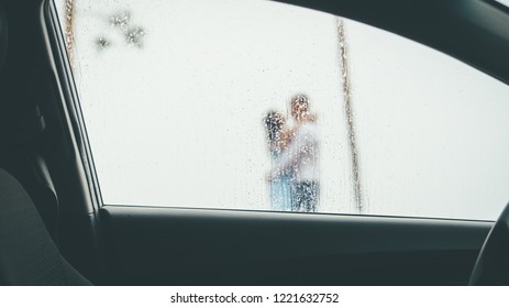 Barely visible silhouettes of groom and bride, standing in each others hugs under the rain, view from inside of a wet moist car