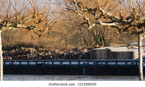 A barge loaded with rubbish or scrap travels along the Main river in the light of the setting sun with bare trees in the foreground