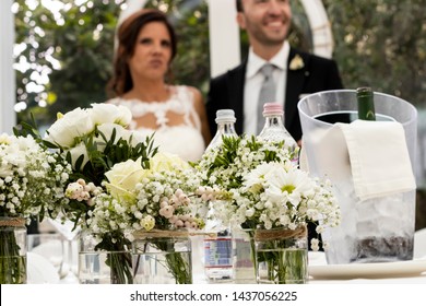 Bari, Italy - 10 06 2018: Wedding In A Farm In Puglia, Region Of Southern Italy. Small Bouquet Of Daisies On The Bride And Groom Table With The Protagonists Of The Wedding In The Background.