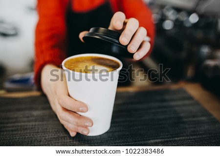 Barista in apron is holding in hands hot cappuccino in white takeaway paper cup. Coffee take away at cafe shop