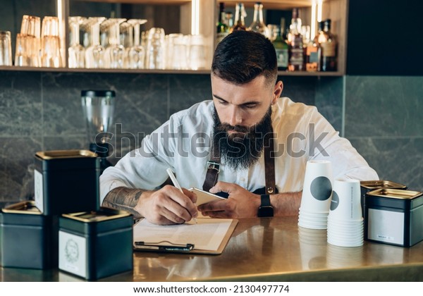 Barista Doing an Inventory of the Products and\
Writing Notes on a Clipboard in a Cafe. \
Serious waiter with a\
beard standing in the bar counter and using mobile phone while\
counting stock tea\
caddy.