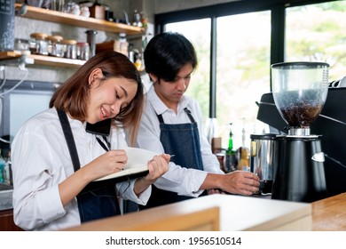 Barista Girl Or Coffee Maker Answer Mobile Phone Also Record The Order Detail Into Notebook While Her Co-worker Is Working With Machine. Concept Of Good Teamwork Support Small Business System.