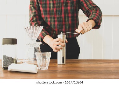 Barista is grinding coffee beans with manual stainless steel grinder to make black coffee machine, brewing equipment or coffee drip set Dripper on a wooden table In the kitchen at home in the morning