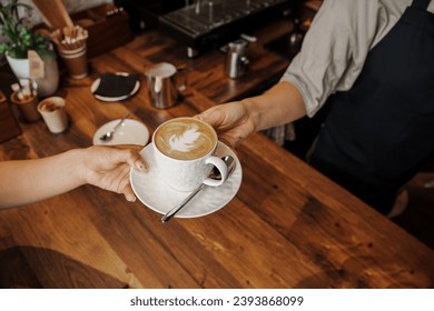 A barista hands over a beautifully crafted cappuccino with latte art to a customer at a wooden cafe counter - Powered by Shutterstock