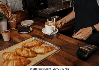 A barista places a cappuccino with latte art next to freshly baked croissants on a wooden counter in a cozy cafe setting - Powered by Shutterstock