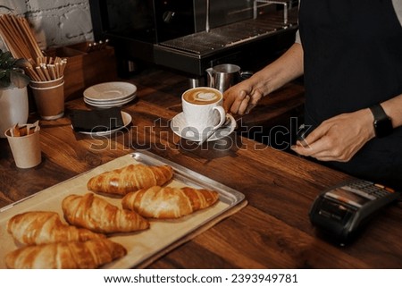 A barista places a cappuccino with latte art next to freshly baked croissants on a wooden counter in a cozy cafe setting [[stock_photo]] © 