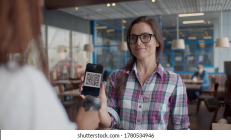 Barista Scanning QR Code In Customer Smartphone At Counter In Cafeteria. Young Woman Client Showing Barcode On Mobile Phone For Discount In Coffee Shop. New Payment Technology Concept.