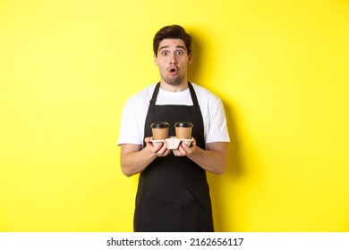 Barista Serving Coffee, Looking Surprised At Camera, Wearing Black Apron, Standing Against Yellow Background