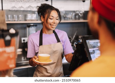 Barista is smiling while taking an order at the counter and holding a yellow cup of coffee, her customer is paying using a digital device
