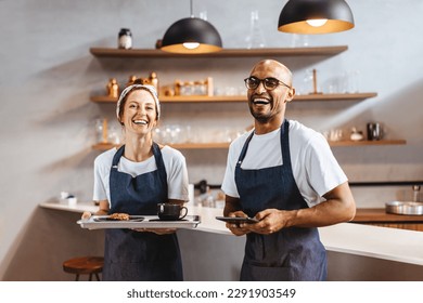 Baristas stand side by side in a cafe, ready to deliver an excellent dining experience to their customers. Two happy waitstaff members holding a tray of food and a tablet in a coffee shop. - Powered by Shutterstock