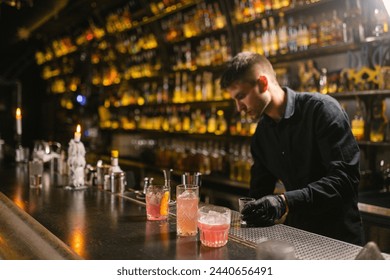 Barkeeper stands behind bar counter with done cocktails for resting visitors of pub. Young worker looks for bottle with alcohol to make new drinks - Powered by Shutterstock