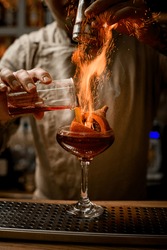 Barman Add Ingredients To Cocktail Glass With Small Orange Pumpkin And Yellow Pepper Inside And Makes Fire Over It