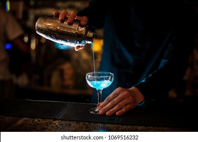 barman in a blue shirt pours from a shaker into a glass of alcohol cocktail Blue Lagoon on a dark background in the bar