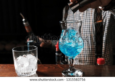 barman in a blue shirt pours from a shaker into a glass of alcohol cocktail Blue Lagoon on a dark background in the bar art