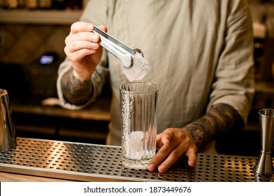 Barman Holding Metal Tongs In Hand With Ice Cube Over Cocktail Glass On Bar Counter Surface