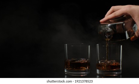 Barman Pour Golden Whiskey, Cognac Or Brandy From Bottle Into Glasses With Ice Cubes On Black Dark Background. Bartender Pouring Tasty Alcohol Cocktail Drink Bourbon, Rum. Copy-space