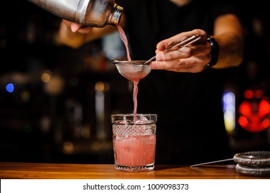 Barman Poured An Alcoholic Cocktail Of Pink Color From The Shaker Through A Strainer Into A Crystal Glass Using Bar Equipment