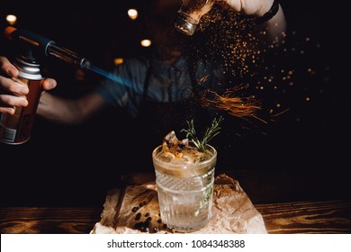 Barman prepares cocktail with orange and herbs in transparent glass on bar with alcohol. Uses burner with sparks. Dark background. - Powered by Shutterstock