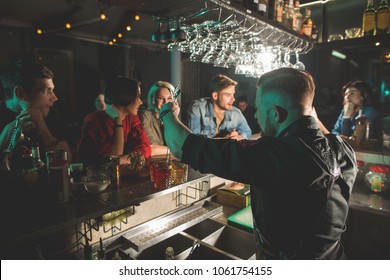 Barman preparing alcoholic drink while standing at counter. Visitors sitting opposite him. Leisure and work in bar concept - Powered by Shutterstock