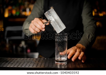 Barman putting a big rectangular piece of ice into a cocktail glass for making a tasty alcoholic drink