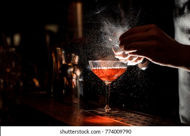 Barman`s hands sprinkling the juice into the cocktail glass filled with alcoholic drink on the dark background
