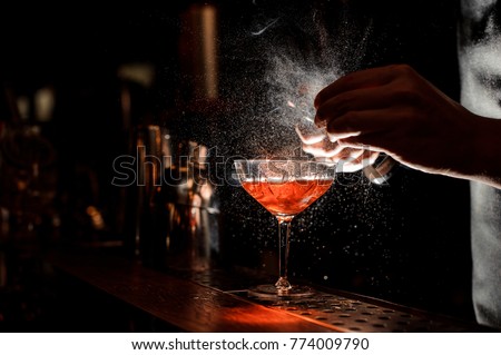 Barman`s hands sprinkling the juice into the cocktail glass filled with alcoholic drink on the dark background