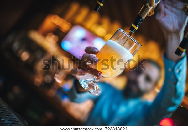 Barmen\
or brewer filling glass with beer. Barmen is pouring lager beer to\
glass from  beer taps. Bar or night club\
interior